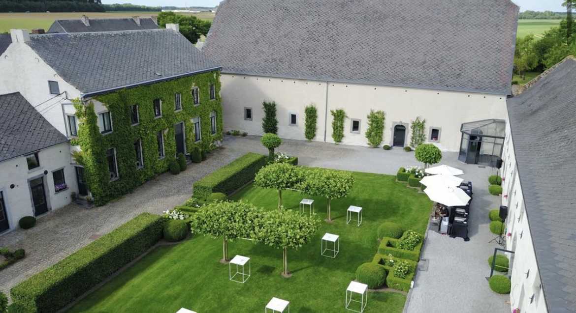 Screenshot Vue aérienne d'une cour du Domaine de Mehaignoul avec pelouse, tables blanches, bâtiments en pierre et façade couverte de lierre.