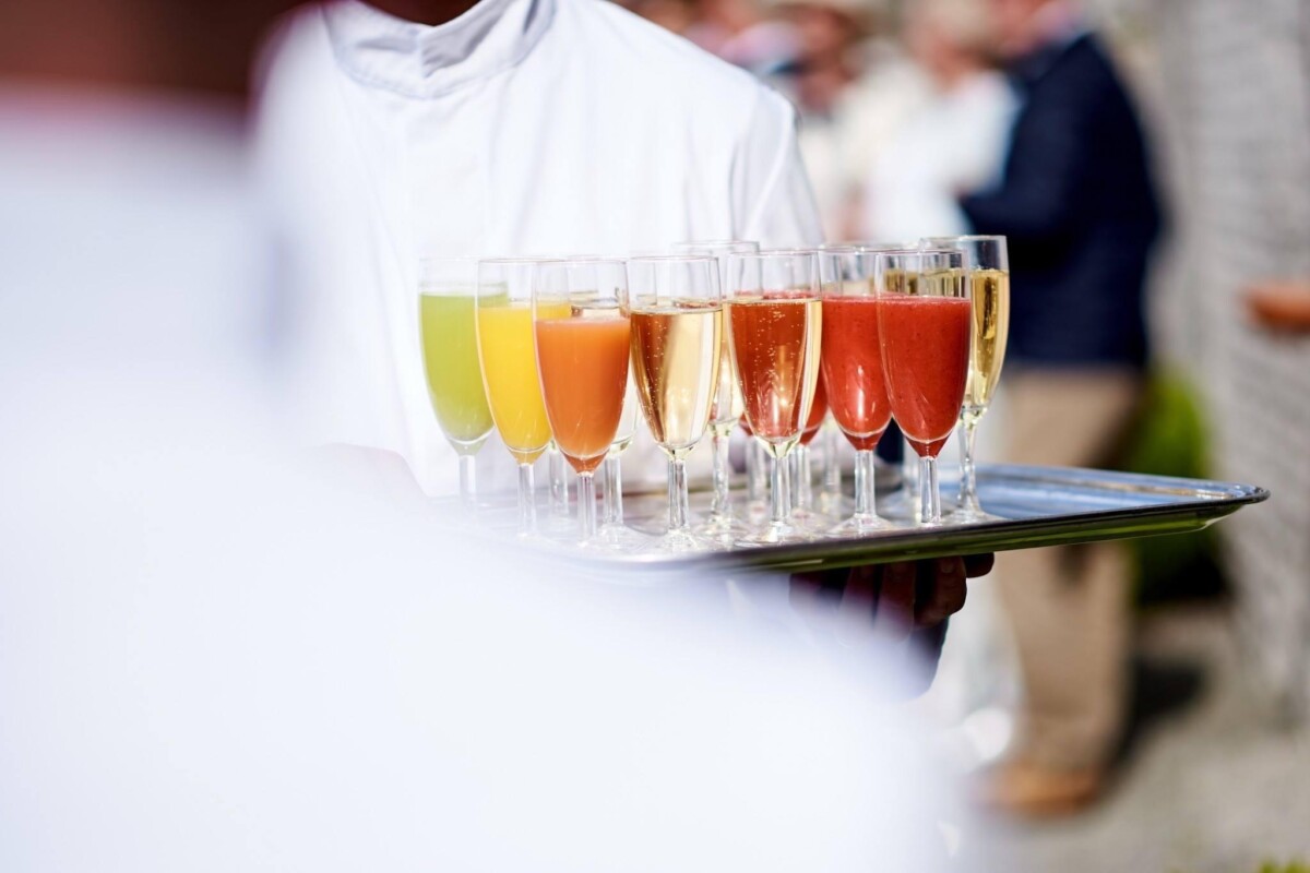 domaine-de-mehaignoul119 Un serveur en uniforme blanc porte un plateau de verres remplis de cocktails colorés, dont des rouges, jaunes et orange.