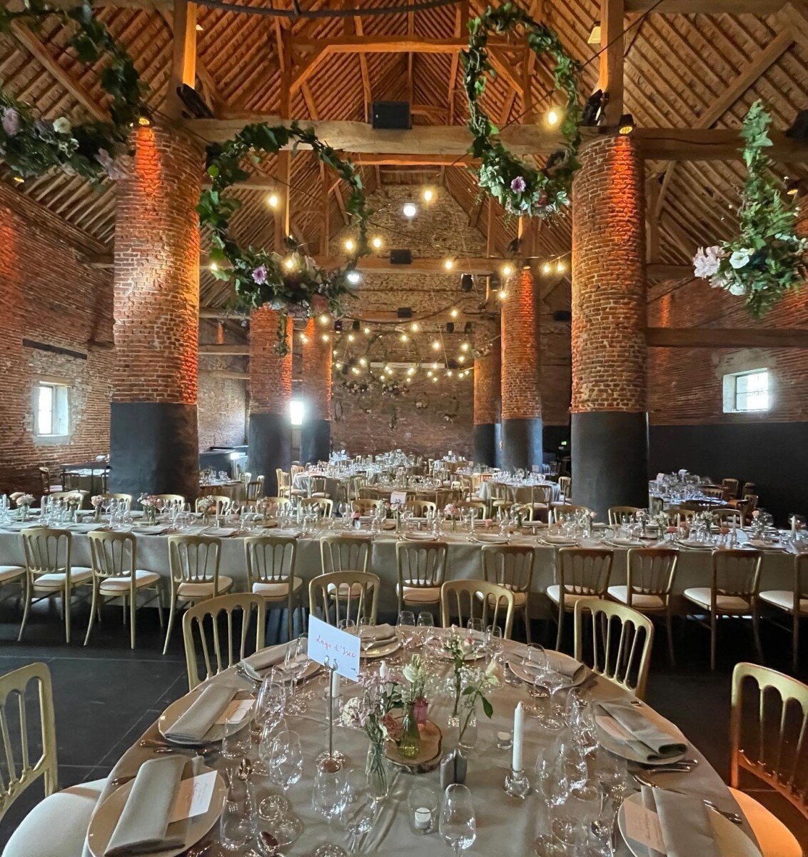 domaine-de-mehaignoul125 Salle de réception en briques avec tables rondes dressées, chaises dorées, décorations florales suspendues et lumières tamisées.