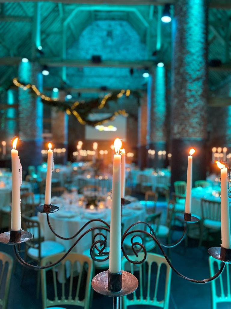 domaine-de-mehaignoul22 Chandelier avec bougies allumées devant des tables rondes dressées dans une salle aux murs en briques et éclairage bleu.