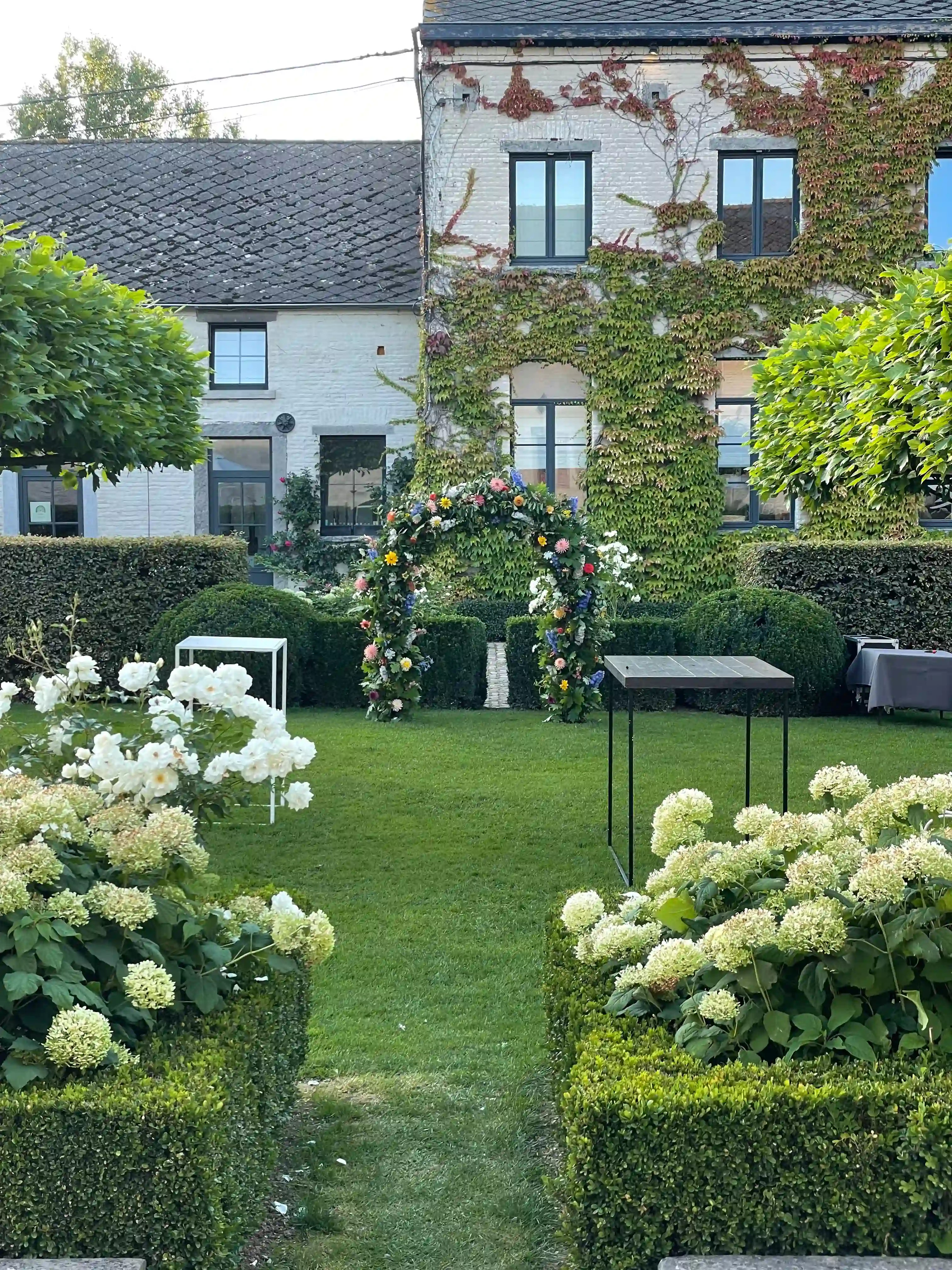 domaine-de-mehaignoul73 (1) Jardin verdoyant avec des hortensias blancs, une arche florale colorée et une façade de maison couverte de lierre.