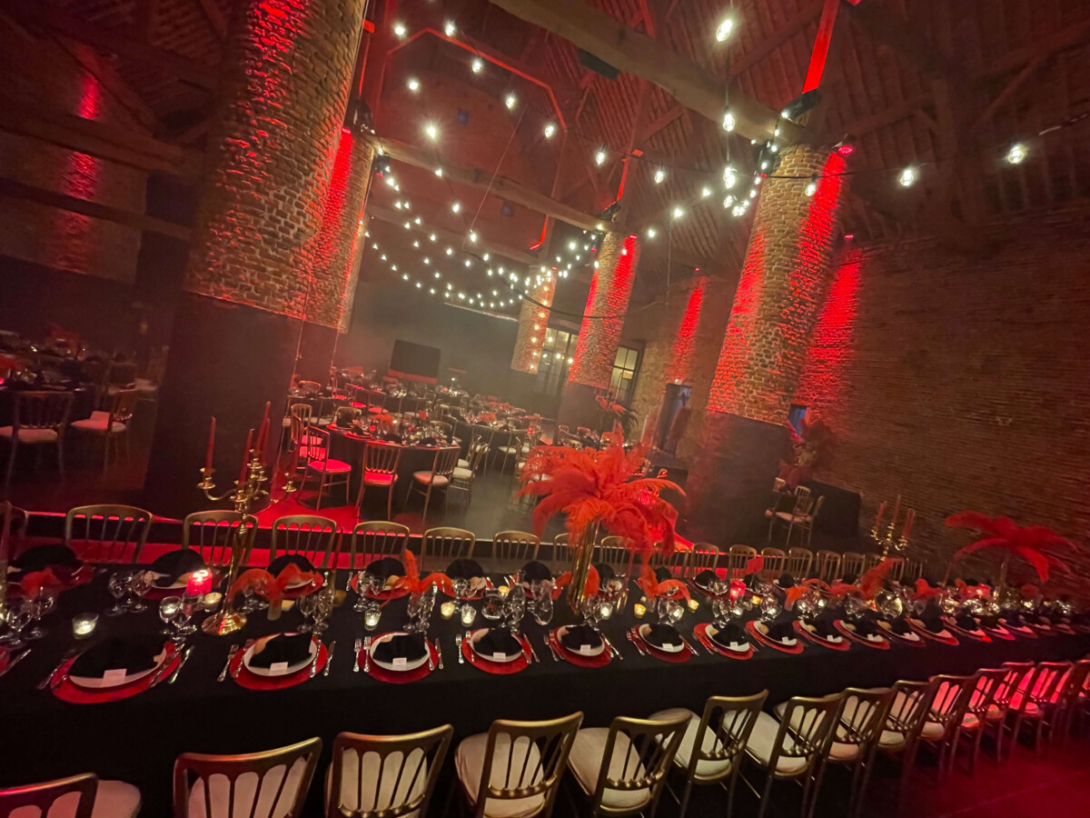 evenement-traiteur-mariage-mehaignoul145 Tables de réception élégamment dressées avec nappes noires, chandeliers dorés et décorations rouges, dans une salle en briques éclairée par des lumières suspendues.