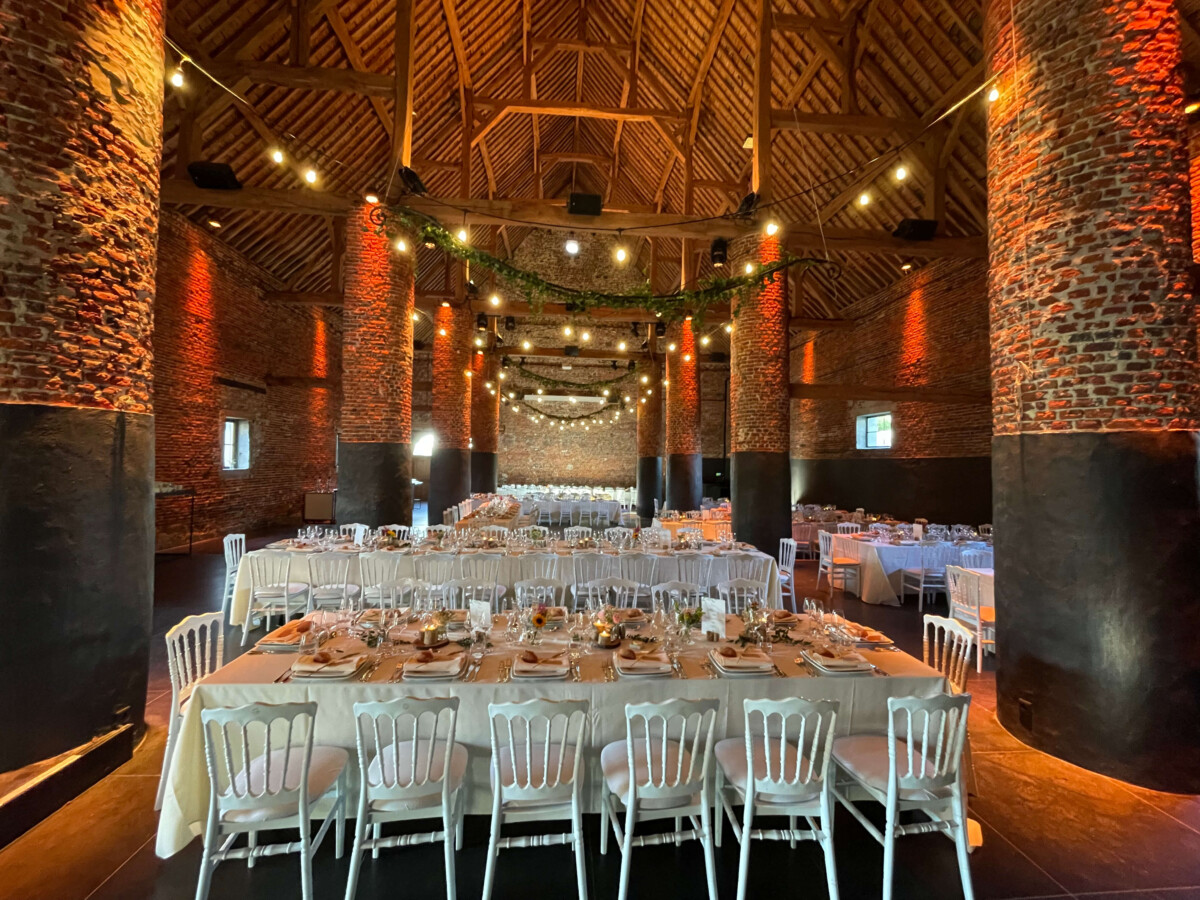 evenement-traiteur-mariage-mehaignoul244 Salle de réception en briques rouges avec tables dressées et chaises blanches, éclairée par des guirlandes lumineuses suspendues au plafond en bois.