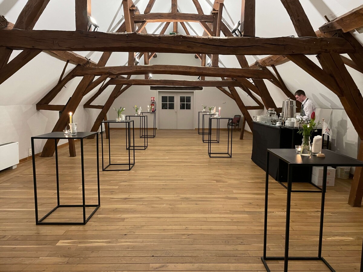 evenement-traiteur-mariage-mehaignoul294 Salle avec poutres apparentes, tables hautes noires avec bougies et fleurs, et un serveur au bar à l'arrière-plan.