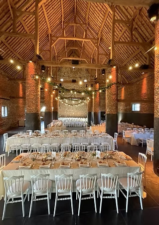 la-grange Des tables décorées avec des nappes beige et des chaises blanches sont disposées dans une grande salle en briques avec un plafond en bois.