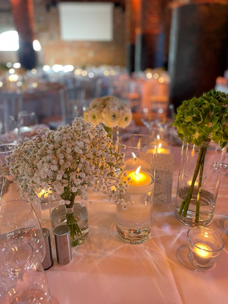 mariage-faq Bouquet de fleurs blanches et vertes sur une table avec bougies allumées, dans une salle en briques rouges décorée pour un événement.