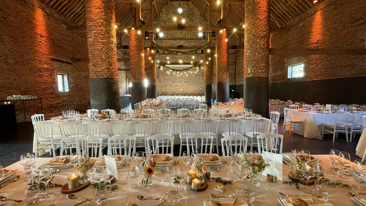mariage-salle Grande salle en briques avec tables dressées, chaises blanches et guirlandes lumineuses, prête pour un événement au Domaine de Mehaignoul.
