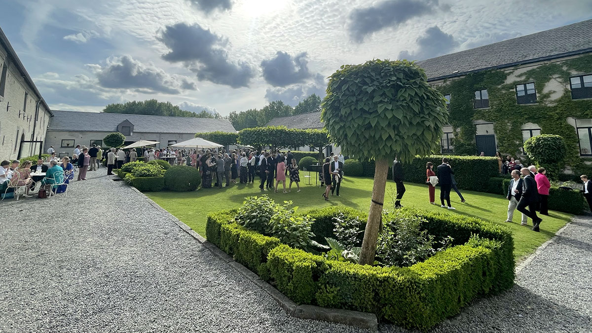 mariage03 Des invités en tenue de cérémonie se rassemblent dans le jardin verdoyant du Domaine de Mehaignoul, entouré de bâtiments en pierre.