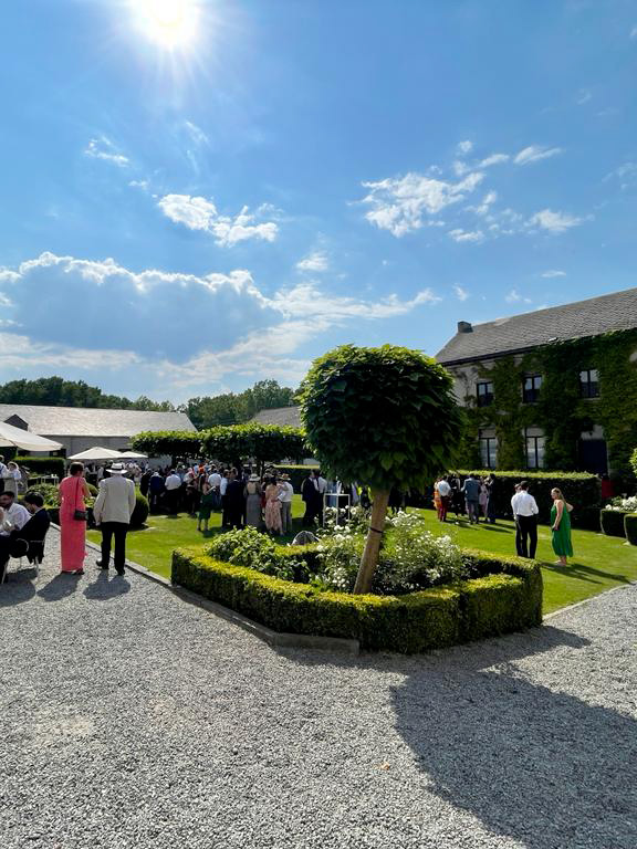 reception-faq Des invités en tenue élégante se rassemblent dans le jardin d’un domaine, entourés de bâtiments en pierre et de végétation luxuriante.