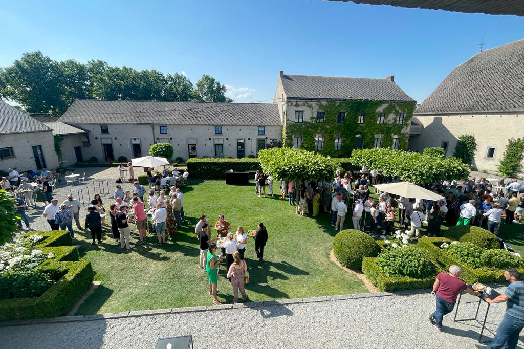 Des invités rassemblés dans la cour d'une ferme en pierre avec toits en ardoise, entourée de haies taillées et de parasols blancs.