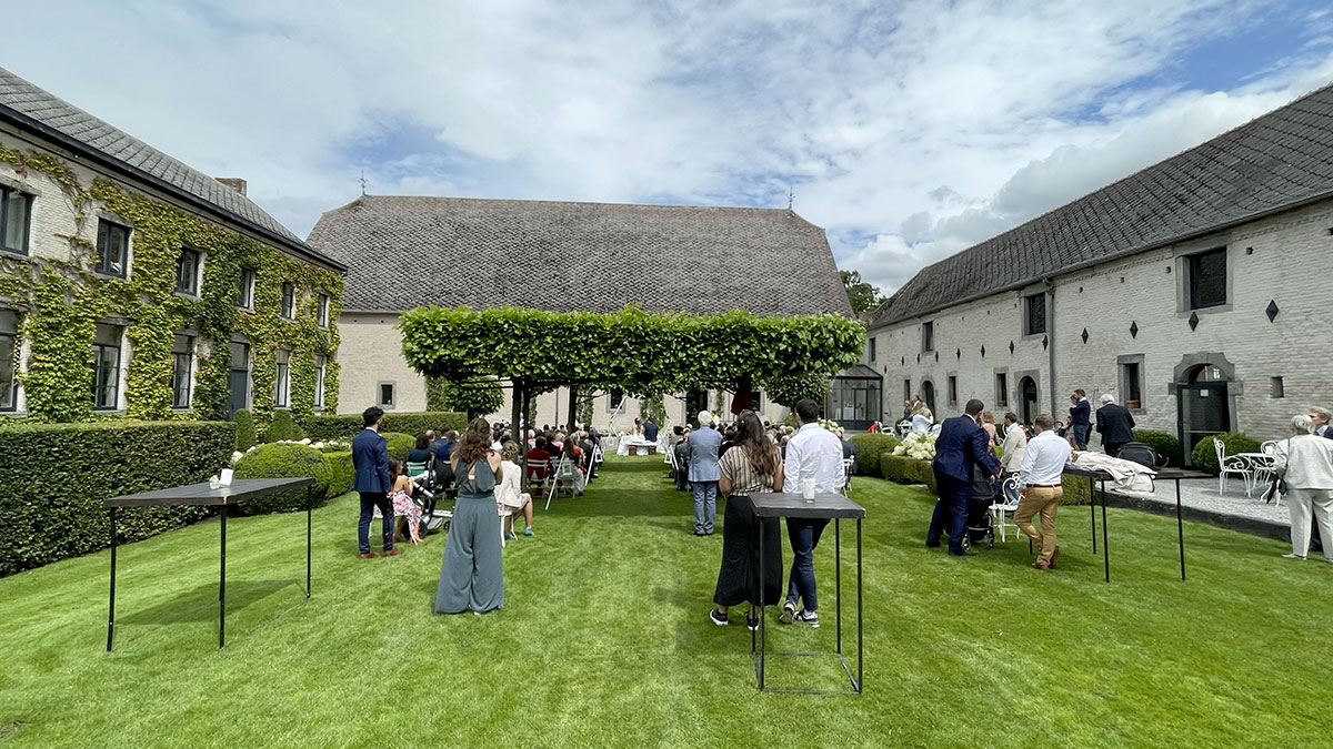 reception-salle Des invités se rassemblent sur une pelouse entourée de bâtiments en pierre, avec des tables hautes et des arbres taillés.