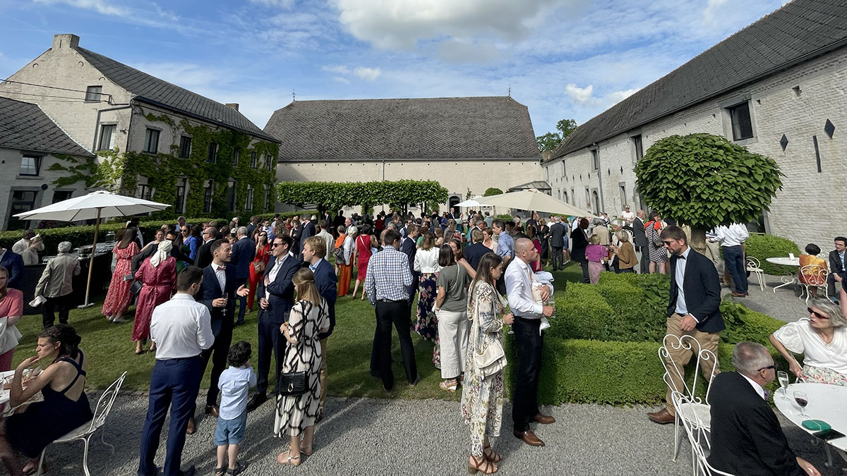 reception01 Des invités en tenue élégante se rassemblent dans la cour d'un domaine en pierre, entourée de bâtiments couverts de lierre et de parasols blancs.