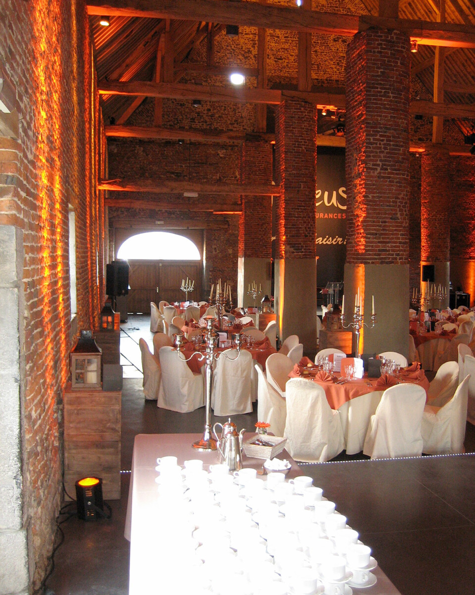 seminaire-hero Salle en briques avec tables rondes dressées, nappes orange, chaises blanches, chandeliers et service à café, sous plafond en bois.