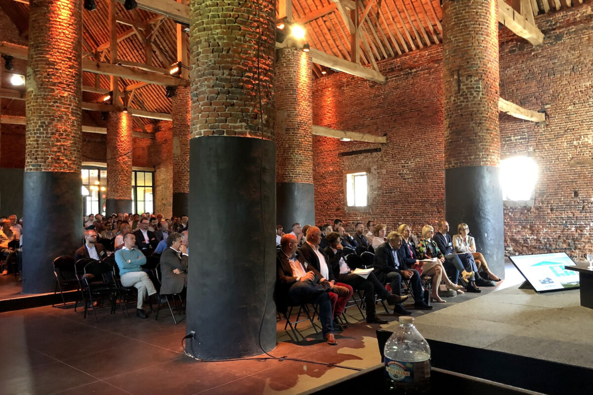 Des personnes assises dans une salle en briques avec de grandes colonnes, écoutant une présentation au Domaine de Mehaignoul.