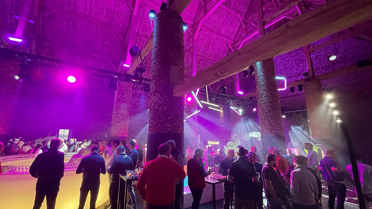 seminaire02 Des invités se rassemblent sous un éclairage violet dans une grande salle avec des piliers en briques et un plafond en bois.